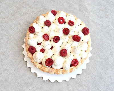A tart covered in piped cream and raspberries, on a scalloped board.
